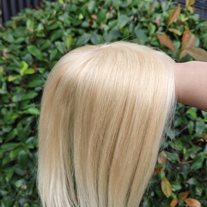 May include: A hair topper featuring straight, light blonde hair with a visible monofilament base at the crown. The hair is smooth and falls in a sleek, layered style, showcasing a natural-looking part and a consistent, bright platinum blonde color.