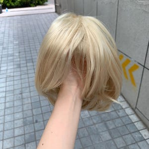 Könnte beinhalten: Eine kurze, gestufte Perücke in einem hellen Platinblond. Das Haar hat eine glatte Textur mit weichen, abgerundeten Enden, die sich nach innen zum Nacken hin wölben. Die Fasern haben einen dezenten Glanz und einen natürlich wirkenden Scheitel.