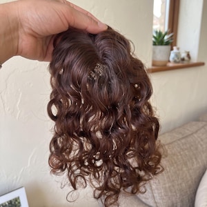 May include: A hair topper piece featuring medium brown, curly hair strands with a defined part. The hair is styled in loose, bouncy ringlets that cascade downwards. The base of the hairpiece includes small, visible metal clips for secure attachment.