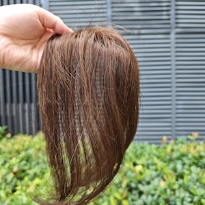 Può includere: Un topper per capelli con ciocche lisce color marrone medio e sottili riflessi castano chiaro. I capelli sono disposti in uno stile liscio e scalato con una struttura di base visibile sulla parte superiore, progettata per offrire copertura e volume.