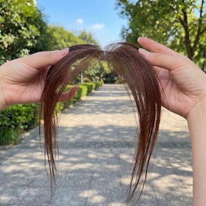 Könnte beinhalten: Ein braunes Haarteil mit einer dünnen, transparenten Basis und langen, glatten Strähnen. Das Haarteil hat einen Mittelscheitel mit feinem, fransigem Pony, der das Gesicht umrahmt. Die Fasern sind in einem mittleren Braunton mit Reflexen.