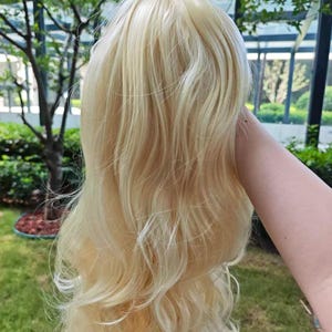 Könnte beinhalten: Eine lange, gewellte Perücke aus hellblondem Haar mit dezenten platinfarbenen Highlights. Das Haar hat eine gestufte, voluminöse Textur mit weichen Locken, die über die Länge fallen und ein natürliches Aussehen mit verschiedenen Blondtönen erzeugen.