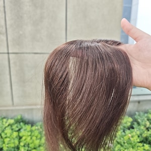 Puede incluir: Una pieza de cabello postizo en color marrón con una base de monofilamento y una raya visible. El cabello es liso, suave y de color marrón medio, diseñado para cubrir la zona de la coronilla con una apariencia natural del cuero cabelludo.