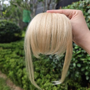 Puede incluir: Una extensión de cabello de clip con cabello rubio claro y liso, peinado como flequillo con secciones laterales más largas. El cabello tiene una textura suave y fina, mostrando un color rubio platino brillante en toda la pieza.