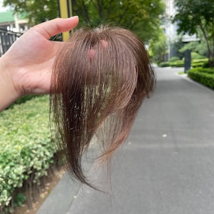 以下が含まれることがあります： 自然な前髪を模した、ストレートでレイヤー状の毛束を持つブラウンのヘアピース。髪は細く滑らかで、明るい茶色と暗い茶色の微妙なグラデーションがあり、ヘアアクセサリーとしてリアルな外観を作り出しています。