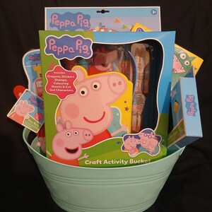 May include: A light blue metal bucket filled with Peppa Pig themed craft activity items. The central box features Peppa Pig, includes crayons, stickers, stamps, colouring sheets, and cut-out characters. Additional Peppa Pig branded items are visible.