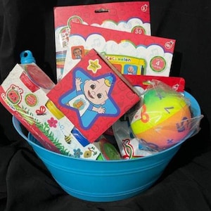 May include: A blue metal basket filled with children's toys and books. Items include a red box with a star design, a colorful ball, and several books with the "Cocomelon" logo. The basket is set against a dark background.