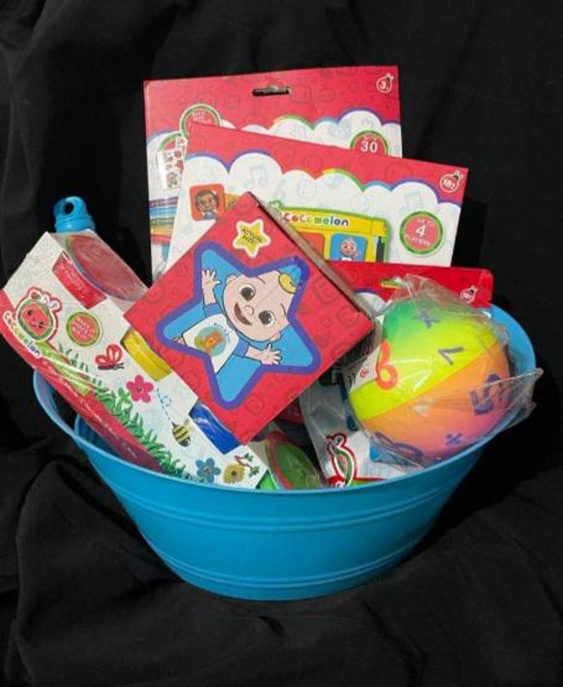 May include: A blue metal basket filled with children's toys and books. Items include a red box with a star design, a colorful ball, and several books with the "Cocomelon" logo. The basket is set against a dark background.