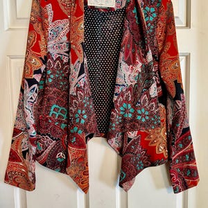 Op de afbeelding: Een levendige rode blazer met een paisley patroon in tinten van teal, oranje en zwart. De open jas heeft een zwarte stippen voering en een label met de tekst "Cartonnier".
