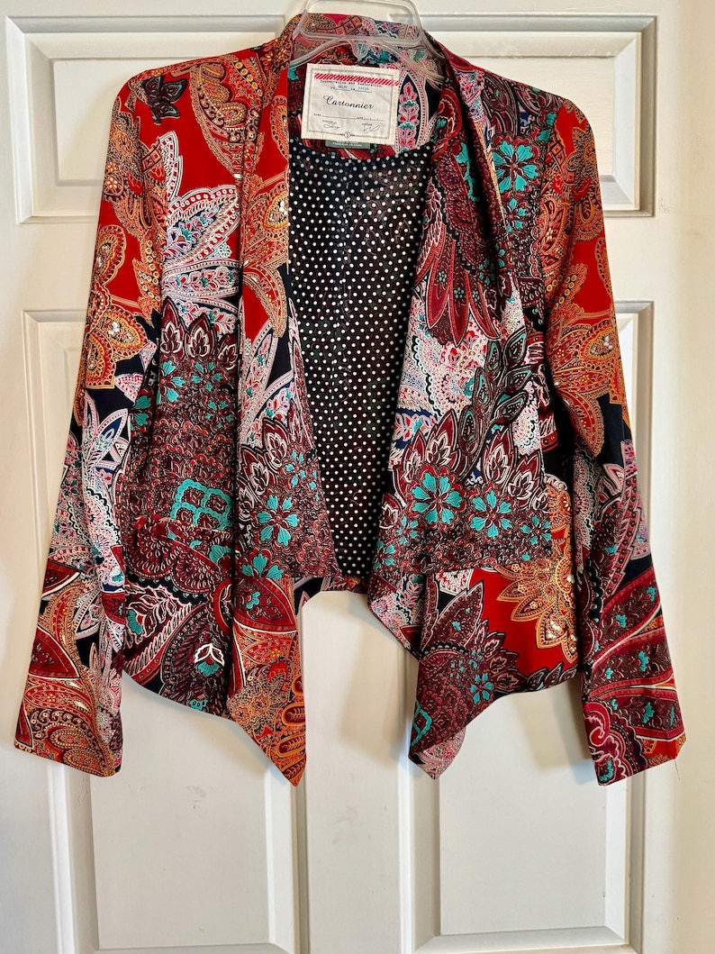 Op de afbeelding: Een levendige rode blazer met een paisley patroon in tinten van teal, oranje en zwart. De open jas heeft een zwarte stippen voering en een label met de tekst "Cartonnier".
