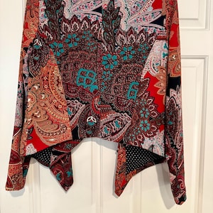 Op de afbeelding: Een levendige, openvallende jas met een opvallend paisley-patroon in tinten rood, bruin en blauwgroen. De jas heeft lange mouwen en een unieke asymmetrische zoom, wat zorgt voor een stijlvolle en artistieke uitstraling.