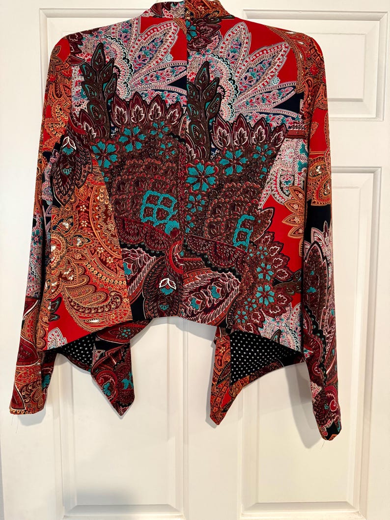 Op de afbeelding: Een levendige, openvallende jas met een opvallend paisley-patroon in tinten rood, bruin en blauwgroen. De jas heeft lange mouwen en een unieke asymmetrische zoom, wat zorgt voor een stijlvolle en artistieke uitstraling.