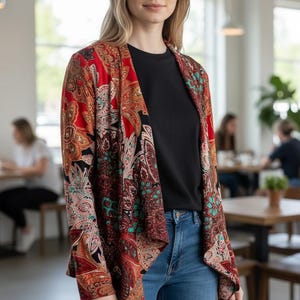 Op de afbeelding: Een vrouw draagt een open vest met rode, oranje en zwarte paisley-print over een zwart t-shirt en een blauwe spijkerbroek. Het vest heeft een gedrapeerde voorkant en lange mouwen. De achtergrond is een caf&eacute; met tafels en stoelen.