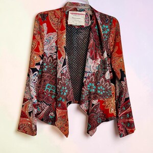 Op de afbeelding: Een kleurrijke open blazer met een paisley patroon in rood, teal en bruin. De blazer heeft een zwarte stippenvoering en een label bij de halslijn. De mouwen zijn lang en de snit is asymmetrisch.