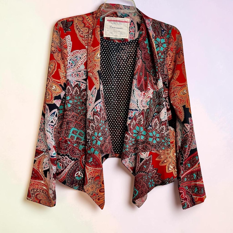 Op de afbeelding: Een kleurrijke open blazer met een paisley patroon in rood, teal en bruin. De blazer heeft een zwarte stippenvoering en een label bij de halslijn. De mouwen zijn lang en de snit is asymmetrisch.