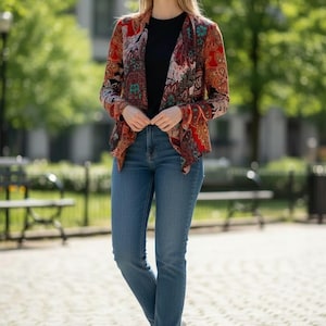 Op de afbeelding: Een vrouw draagt een jasje met patroon in rood, oranje en turquoise accenten over een zwarte top en een blauwe jeans. Ze draagt witte sneakers. De achtergrond is een park met bomen en banken.