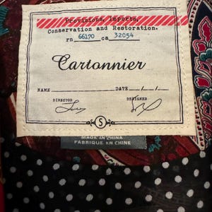 Op de afbeelding: Close-up van een kledinglabel met de merknaam "Cartonnier". Het label bevat de tekst: "Provisions, Imports, Conservation and Restoration." Het label is genaaid op een patroonstof. Het label zegt ook "MADE IN CHINA FABRIQUE EN CHINE."