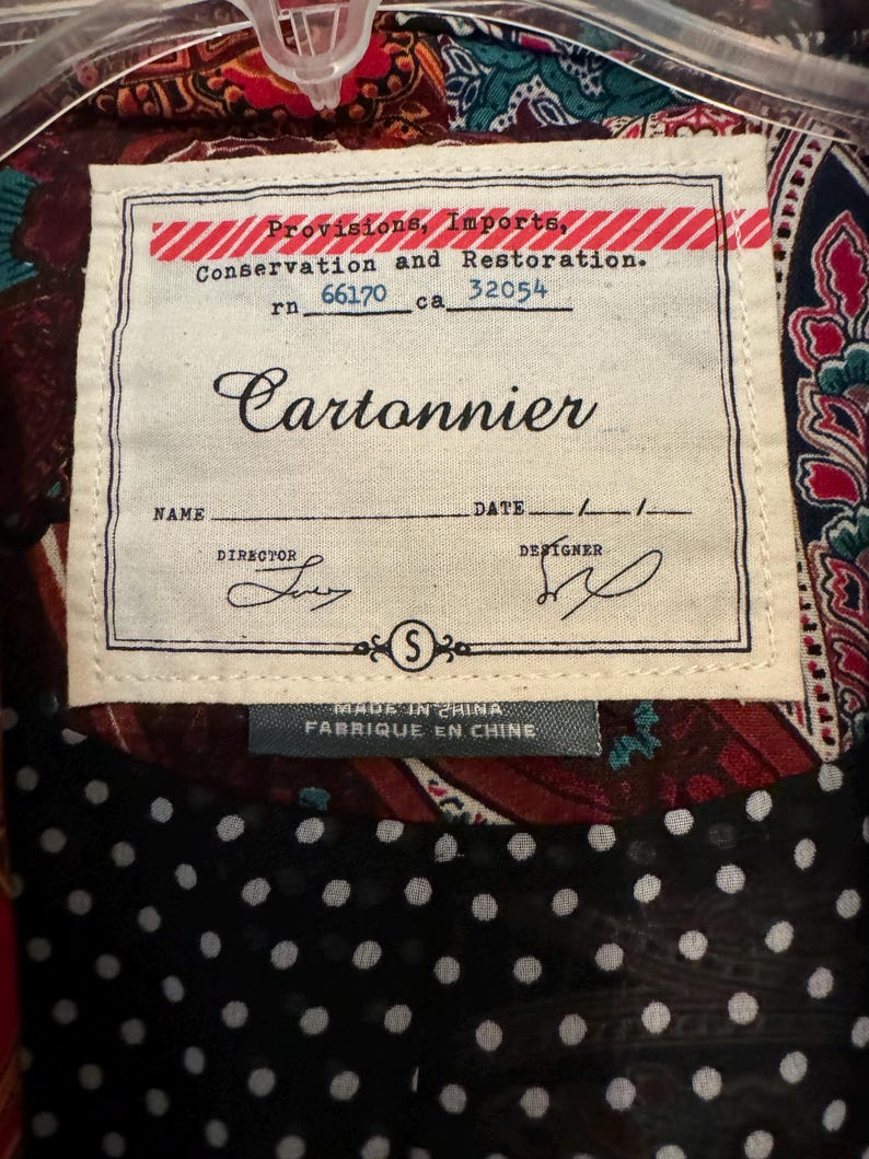 Op de afbeelding: Close-up van een kledinglabel met de merknaam "Cartonnier". Het label bevat de tekst: "Provisions, Imports, Conservation and Restoration." Het label is genaaid op een patroonstof. Het label zegt ook "MADE IN CHINA FABRIQUE EN CHINE."