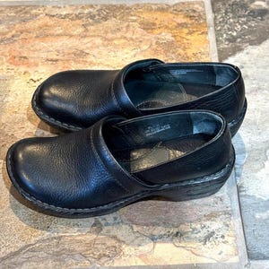 May include: A pair of black leather clogs with a rounded toe and a low heel. The clogs have a slip-on design with a strap across the top of the foot. The shoes are sitting on a stone tile surface.