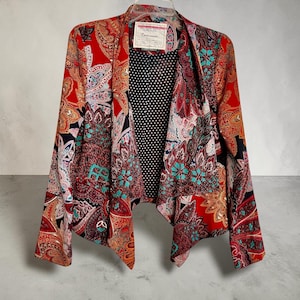 Op de afbeelding: Een levendige blazer met open voorkant en een kleurrijk paisley-patroon in rood, oranje, teal en zwart. De blazer heeft een zwarte stippenvoering en een label bij de halslijn. Het kledingstuk hangt aan een witte hanger.