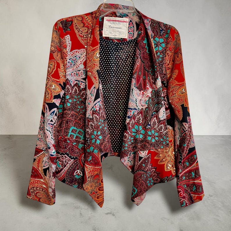 Op de afbeelding: Een levendige blazer met open voorkant en een kleurrijk paisley-patroon in rood, oranje, teal en zwart. De blazer heeft een zwarte stippenvoering en een label bij de halslijn. Het kledingstuk hangt aan een witte hanger.