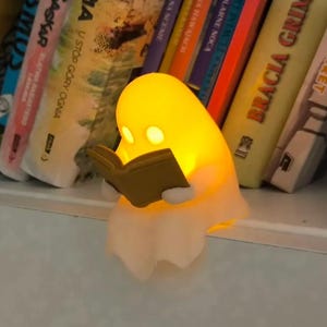May include: A small, glowing, yellow ghost-shaped lamp made of smooth material, featuring two circular eye cutouts and holding a small, solid brown book in its hands. The lamp emits a warm, soft light from within its translucent, rounded body.