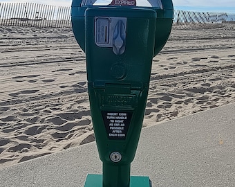 May include: A dark green parking meter with a circular top displaying "SIXTY MIN. TIME LIMIT" and "TIME EXPIRED." The meter has a coin slot and instructions to insert coins and turn the handle. It stands on a square base on a concrete surface, with a beach and fence in the background.