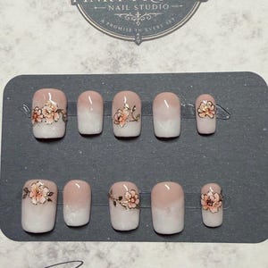 May include: A set of ten press-on nails featuring a nude to white ombre gradient finish. Several nails are decorated with delicate hand-painted peach and gold floral accents. The nails are displayed on a dark gray card with the text Pinky Promise Nail Studio.