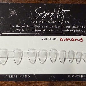 Op de afbeelding: Een maattabelset voor plaknagels met een zwart-wit marmeren kaart met de tekst Sizing Kit for press-on nails. De kaart toont tien doorzichtige, amandelvormige plastic nageltips die in een rij op een transparante strip zijn geplaatst.