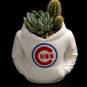 Peut inclure: Un pot de fleurs blanc en forme de sweat-shirt à capuche arborant un logo circulaire rouge et bleu avec le texte CUBS au centre. Le pot contient une plante succulente verte et deux petits cactus aux épines jaunes, sur fond noir.
