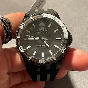 May include: A black Invicta watch with a round face, featuring a black dial with white hour markers, silver hands, and a rotating bezel with white numerals. The dial displays the text Invicta and Water Resis 100M. It has a black silicone strap.