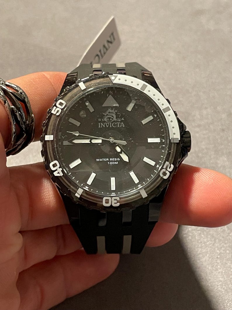 May include: A black Invicta watch with a round face, featuring a black dial with white hour markers, silver hands, and a rotating bezel with white numerals. The dial displays the text Invicta and Water Resis 100M. It has a black silicone strap.