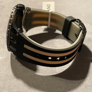 May include: A wristwatch with a black and beige silicone strap featuring two parallel beige stripes running along the length of the band. The watch case is black with a textured bezel, and a small white tag with the text INV is attached to the strap.