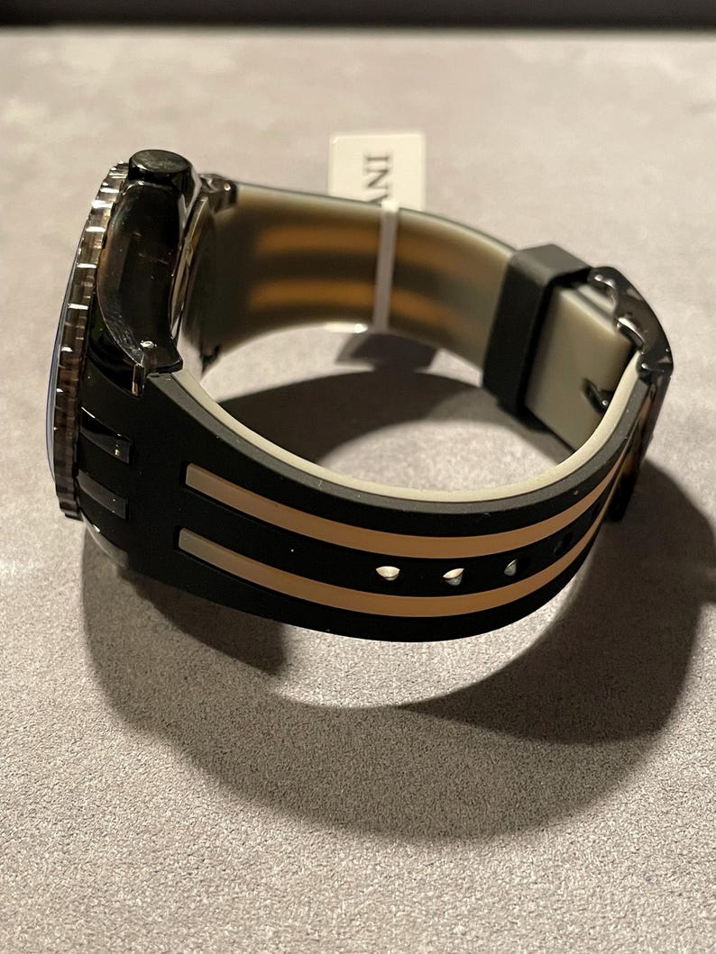 May include: A wristwatch with a black and beige silicone strap featuring two parallel beige stripes running along the length of the band. The watch case is black with a textured bezel, and a small white tag with the text INV is attached to the strap.