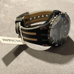 May include: An Invicta wristwatch featuring a black dial and a black and translucent silicone strap. The watch has a rotating bezel with white numerals and a white tag attached to the band that displays the brand name Invicta in black text.