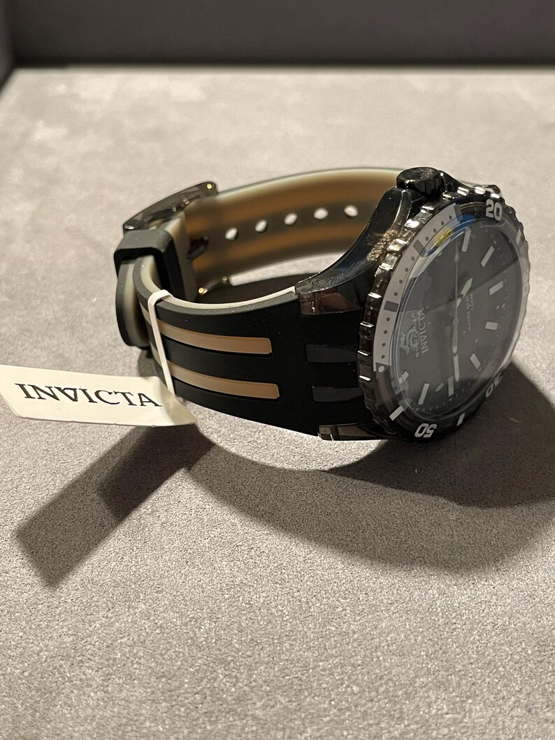 May include: An Invicta wristwatch featuring a black dial and a black and translucent silicone strap. The watch has a rotating bezel with white numerals and a white tag attached to the band that displays the brand name Invicta in black text.