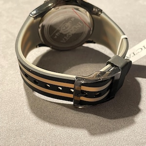 May include: A wristwatch with a translucent grey case and a black and beige striped silicone strap. The watch features a round silver-tone case back with the Invicta logo engraved in the center. A black buckle secures the strap, and a white tag is attached.