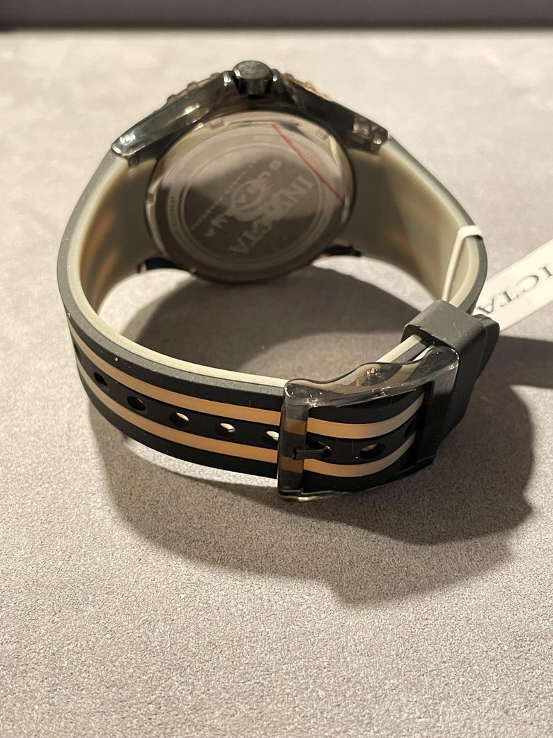 May include: A wristwatch with a translucent grey case and a black and beige striped silicone strap. The watch features a round silver-tone case back with the Invicta logo engraved in the center. A black buckle secures the strap, and a white tag is attached.