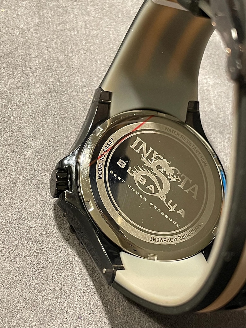 May include: The back of an Invicta Subaqua watch with a black case and a two-tone strap. The stainless steel case back features the text Invicta, Subaqua, Model No. 69447, Water Resistant 100M, Best Under Pressure, and Singapore Movement.