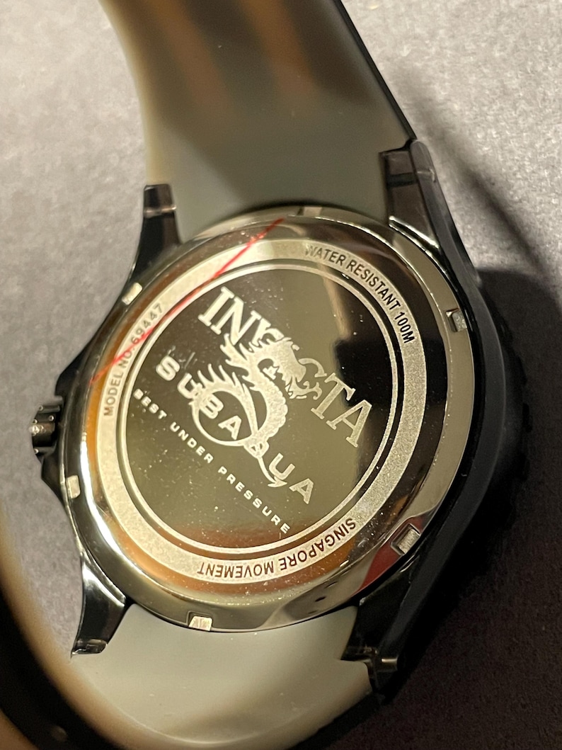 May include: The stainless steel case back of an Invicta Subaqua watch features the text INVICTA SUBAQUA, BEST UNDER PRESSURE, WATER RESISTANT 100M, MODEL NO 69447, and SINGAPORE MOVEMENT. A dragon graphic is engraved in the center of the metallic surface.