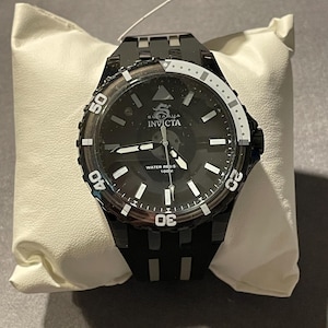 May include: A black Invicta wristwatch featuring a round black dial with white hour markers, a dragon logo, and the text Invicta and Water Resistant 100M. The watch has a black bezel with white numerals and a black silicone strap with silver accents.
