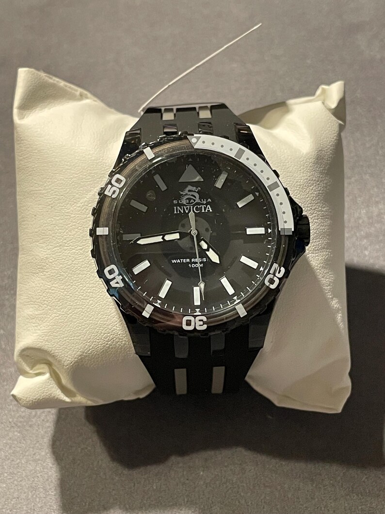 May include: A black Invicta wristwatch featuring a round black dial with white hour markers, a dragon logo, and the text Invicta and Water Resistant 100M. The watch has a black bezel with white numerals and a black silicone strap with silver accents.