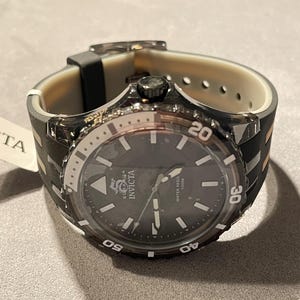 May include: An Invicta watch with a dark gray dial, luminous hands, and markers. The bezel features white numerals 20, 30, 40, and 50. It has a translucent dark gray case and a two-tone black and beige silicone strap with a buckle closure.