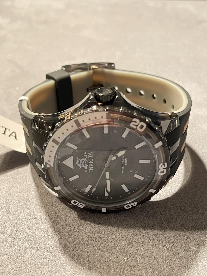 May include: An Invicta watch with a dark gray dial, luminous hands, and markers. The bezel features white numerals 20, 30, 40, and 50. It has a translucent dark gray case and a two-tone black and beige silicone strap with a buckle closure.