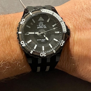 May include: A black Invicta Subaqua watch with a round black dial, luminous hour markers, and silver-tone hands. The bezel features white numerals 20, 30, 40, and 50. The watch includes a black silicone strap and is marked Water Resistant 100M.