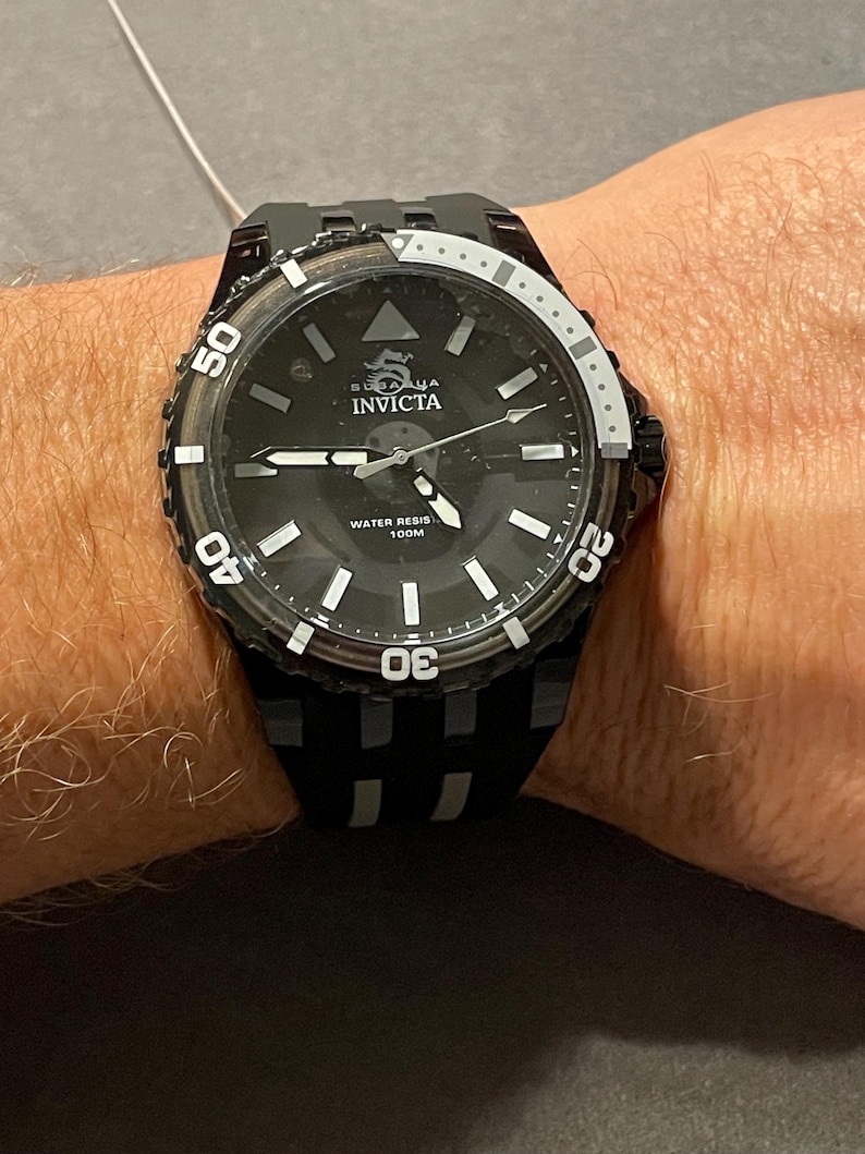 May include: A black Invicta Subaqua watch with a round black dial, luminous hour markers, and silver-tone hands. The bezel features white numerals 20, 30, 40, and 50. The watch includes a black silicone strap and is marked Water Resistant 100M.