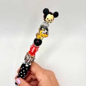 May include: A decorative pen featuring a series of stacked beads, including a Mickey Mouse character topper, a Pluto character charm, and various round beads in black, red, and silver tones, finished with a black and white polka dot patterned base.
