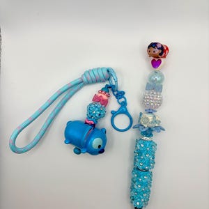 May include: A blue Stitch-themed keychain and pen set. The keychain features a blue Stitch figure and a blue and pink cord. The pen is adorned with blue floral accents and sparkling embellishments, with a black tip.