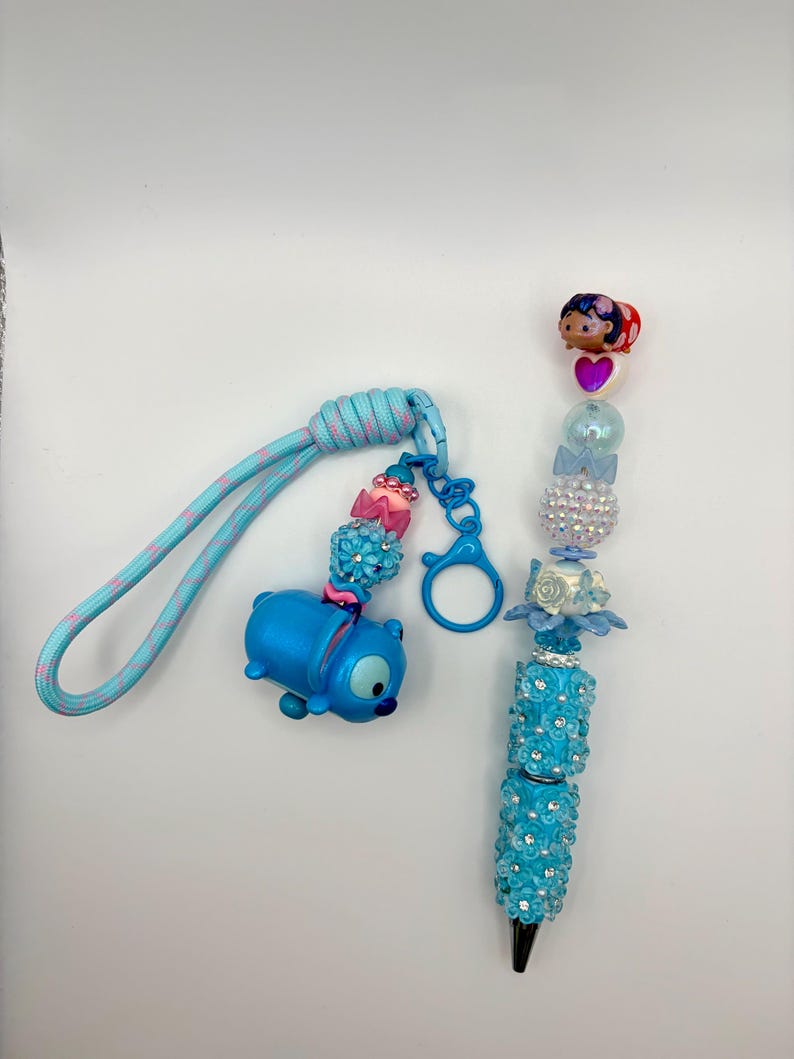 May include: A blue Stitch-themed keychain and pen set. The keychain features a blue Stitch figure and a blue and pink cord. The pen is adorned with blue floral accents and sparkling embellishments, with a black tip.