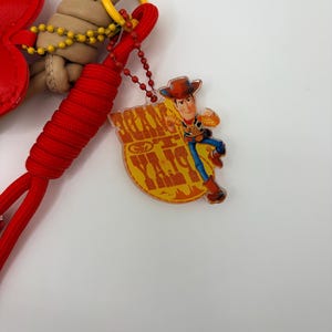 Pu&ograve; includere: Un portachiavi con un cordino intrecciato rosso, una corda annodata color marrone chiaro e un ciondolo a forma di cuore rosso. &Egrave; presente un pendente in acrilico trasparente che raffigura il personaggio Woody di Toy Story con la scritta "TOY STORY" in giallo.